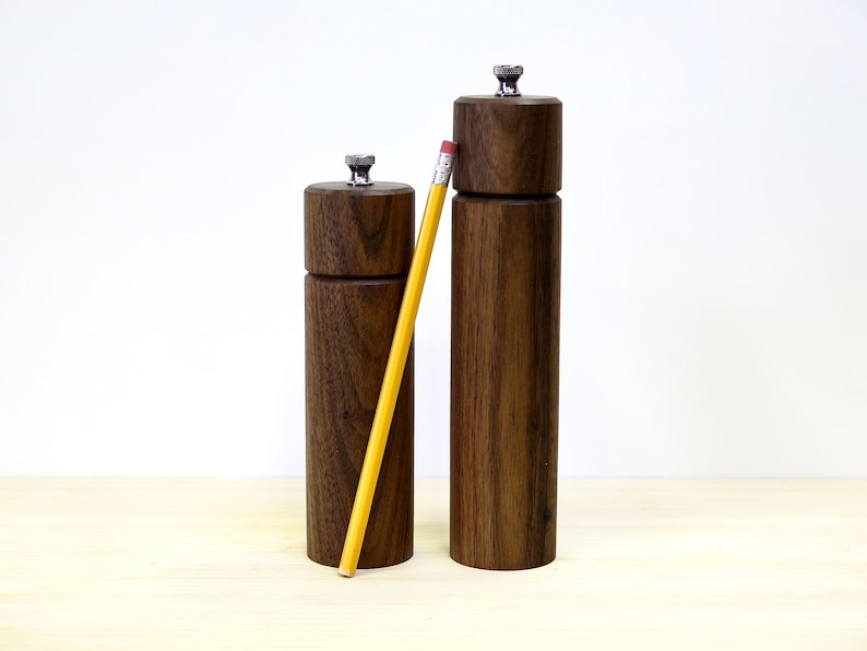 May include: Two wooden salt and pepper grinders with a pencil for scale. The grinders are made of dark brown wood with a cylindrical shape and a silver-colored top. The pencil is yellow with a pink eraser.
