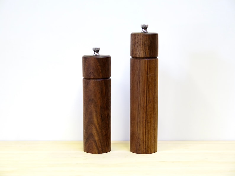 May include: Two wooden salt and pepper grinders. The grinders are made of dark brown wood with a silver-colored top. The grinders are cylindrical in shape and vary in height.