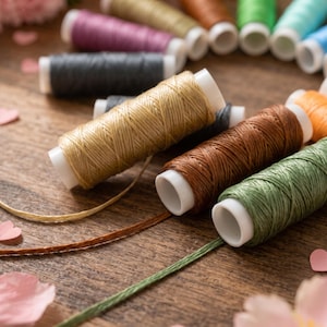 Può includere: Una collezione di rocchetti di filo multicolore disposti in un motivo circolare. I rocchetti presentano varie tonalità tra cui beige, marrone, verde, grigio, viola e arancione, ciascuno avvolto su un nucleo di plastica bianca con fili sciolti.