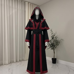 May include: A floor-length black robe featuring deep red trim along the hem, sleeves, and front panels. The garment includes a hooded capelet layer with two circular silver-toned clasps at the shoulders and a wide black sash tied at the waist.