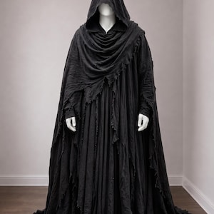 May include: A floor-length black hooded cloak featuring a draped cowl neckline and distressed, frayed edges. The garment is layered over a long, pleated black tunic with fitted sleeves, creating a voluminous silhouette that reaches the floor.