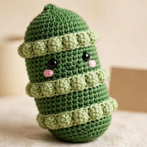 Puede incluir: Un juguete de peluche tejido a ganchillo en color verde con forma de vaina y tres filas horizontales de puntos de burbuja texturizados. El juguete tiene dos ojos de seguridad de plástico negro, una boca pequeña bordada y dos parches rosados.