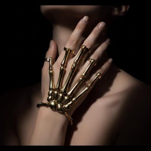 Könnte beinhalten: Ein goldfarbenes Handarmband aus Metall mit skelettartigem Design, das den Handrücken bedeckt und in einzelne Fingerringe übergeht. Das Schmuckstück besteht aus beweglichen Gliedern, die menschlichen Fingerknochen ähneln, und einem Armband.