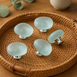 May include: Five oval-shaped cabinet knobs featuring smooth, frosted light blue glass tops and polished silver-toned metal bases. Each knob has a rounded, pebble-like profile, providing a consistent look for furniture hardware or drawer pulls.