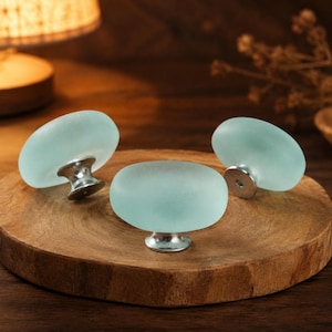 Puede incluir: Tres pomos redondos para armarios con un acabado de vidrio esmerilado en color verde espuma de mar pálido y una base de metal plateado pulido. Cada pomo presenta una superficie superior lisa y redondeada con un vástago metálico.