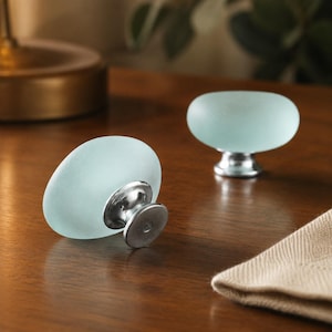 Peut inclure: Deux boutons de placard de forme ovale dotés d'un dessus en verre dépoli bleu clair lisse et d'une base en métal argenté poli. Les boutons présentent un profil arrondi et aplati, conçus pour être fixés sur des meubles ou des tiroirs.