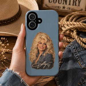 Könnte beinhalten: Eine schieferblaue Smartphone-Hülle mit einem Grafikdruck einer blonden Frau mit voluminösem Haar, die ein Jeanshemd trägt. Der Name Dolly ist in goldener Schreibschrift mit einem kleinen Herzsymbol unter dem Porträt auf der Hülle abgebildet.