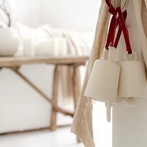 May include: Two off-white ceramic bells suspended by thin red velvet ribbons from a large, tied beige linen fabric bow. The bells have a smooth, matte finish and a simple, tapered shape, hanging together as a decorative ornament.