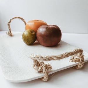 May include: A white rectangular serving tray with rope handles, holding three round fruits. The fruits are a mix of colors: orange, green, and dark red. The tray has a textured surface and is set against a white background.