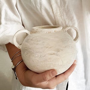 May include: A cream-colored, round ceramic vase with two handles. The vase has a textured surface with a marbled effect. The person holding the vase is wearing a white button-down shirt and silver bracelets.