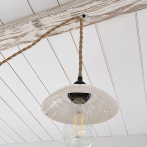 May include: A pendant light fixture featuring a glossy off-white ceramic dome shade suspended by a thick, twisted brown jute rope cord. The fixture includes a black socket housing and a clear glass LED light bulb with visible vertical filaments.