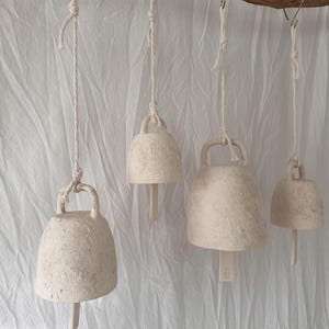 May include: Four off-white ceramic bells with textured surfaces hang from a horizontal wooden branch by white rope cords. Each bell features a top handle and a central clapper. The bells vary in size, and one clapper displays a small embossed logo.
