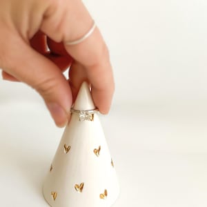 May include: A white ceramic cone-shaped ring holder featuring a pattern of small, metallic gold-colored heart shapes. A silver-toned ring with a square-cut clear stone is positioned near the top point of the cone-shaped display stand.