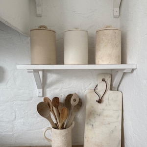 May include: Three cylindrical ceramic canisters with lids sit on a white shelf. Below, a textured ceramic pitcher holds several wooden spoons. Beside the pitcher stands a rectangular white marble cutting board with a brown leather hanging loop.