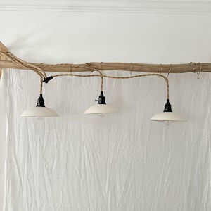 May include: A hanging light fixture featuring three off-white dome-shaped metal shades suspended from a long piece of natural driftwood. Each shade is attached to a black socket and a twisted tan fabric cord, which is wrapped around the wood.