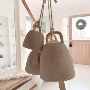 May include: Three off-white, textured ceramic bells of varying sizes suspended by thick, knotted white rope. Each bell features a simple arched handle at the top and a flat, rectangular clapper hanging from the center of the hollow interior.