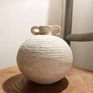 May include: A spherical off-white ceramic vase with a textured, horizontally ribbed surface. The vase features a narrow neck and two small, rounded handles on opposite sides of the opening, creating a balanced and symmetrical appearance.