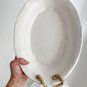 May include: An oval-shaped white ceramic tray with a textured surface. The tray features two sets of thick, natural-colored jute rope handles attached through holes at the top and bottom ends of the piece, providing a sturdy grip for carrying.