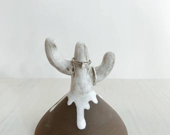 May include: A ceramic ring holder shaped like a cactus with a brown conical base and a white glazed top section. The cactus has two arms extending upwards, with two silver-toned rings resting on the central stem and one of the side arms.