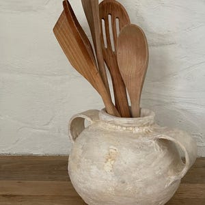 May include: A round, off-white ceramic vessel with two side handles containing a collection of four wooden kitchen utensils, including a solid spoon, a slotted turner, and a flat-edged spatula, all featuring natural wood grain textures.