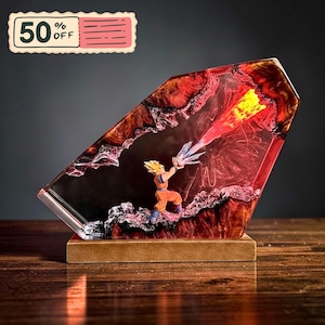 May include: A resin lamp featuring a miniature figure of a spiky-haired character in an orange gi, positioned within a translucent, angular block. The figure is emitting a bright yellow and red energy beam against a textured, dark red background.