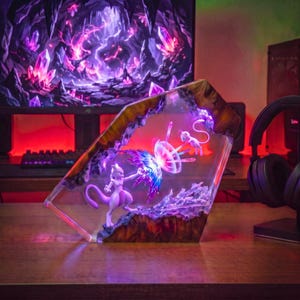 Może przedstawiać: Niestandardowa lampa dioramowa z żywicy przedstawiająca dwie małe fioletowo-białe figurki, Mewtwo i Mew, walczące ze sobą z efektem świetlnym. Scena jest zamknięta w przezroczystym, kanciastym bloku żywicy z ciemną, drewnopodobną obwódką.
