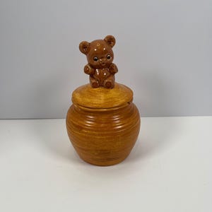 May include: A honey pot made of glazed ceramic with a golden-brown finish and horizontal ribbed texture. The lid features a sculpted brown bear figurine sitting on top, serving as a handle. The pot has a rounded body and a matching circular lid.