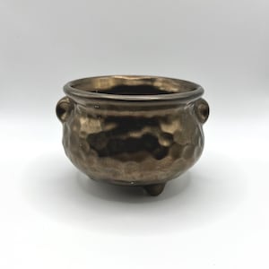 Può includere: Un piccolo calderone in ceramica color bronzo metallizzato con una texture martellata e irregolare. Il recipiente presenta un'ampia apertura, due piccole maniglie laterali e tre piedini corti e arrotondati alla base.