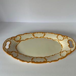 May include: An oval-shaped serving tray featuring a cream-colored center and a raised, textured border in a vibrant mustard yellow. The tray includes two integrated handles on the sides and a subtle grid pattern embossed on the rim area.