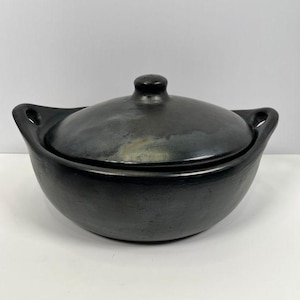 May include: A round, black ceramic cooking pot with a matching lid. The pot features two integrated side handles and a central knob on the lid. The surface has a smooth, dark finish with subtle variations in tone across the rounded body.