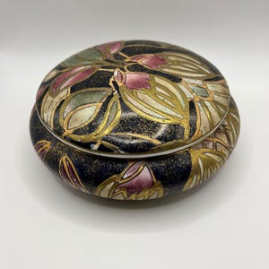 May include: A round ceramic lidded box featuring a dark navy blue background with a speckled gold finish. The surface is decorated with stylized floral motifs in shades of muted pink, sage green, and cream, outlined with gold paint.