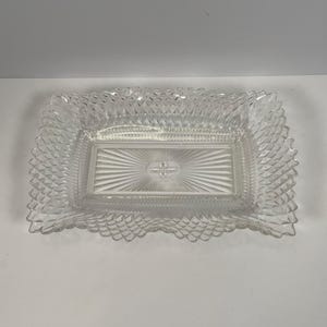 May include: A rectangular clear glass serving tray featuring a central sunburst pattern surrounded by a textured diamond-cut border. The edges of the tray have a scalloped design, and the glass surface is transparent with reflective facets.