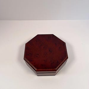 May include: An octagonal wooden box featuring a rich, dark reddish-brown finish with a distinct burl wood grain pattern. The lid is flat with a smooth, polished surface, and the sides of the box are straight, forming a symmetrical eight-sided shape.