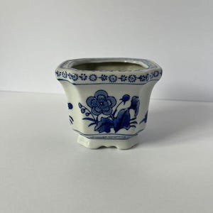 Puede incluir: Una maceta hexagonal de cerámica con base blanca y patrones florales azules. El borde está decorado con un motivo repetitivo de flores pequeñas, mientras que los lados muestran flores y hojas azules más grandes. La maceta tiene un borde acampanado.