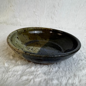 May include: A handmade ceramic bowl with a dark, glossy finish. The bowl features a blend of green, brown, and blue hues, with speckled detailing. The interior is a darker shade than the exterior, creating a contrast. The bowl is suitable for serving food.