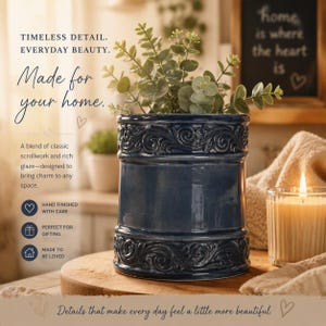 May include: A dark blue ceramic planter featuring embossed scrollwork patterns along the top and bottom rims. The cylindrical vessel contains a green leafy plant and is positioned on a wooden surface next to a lit candle in a glass holder.