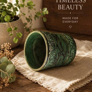 May include: A cylindrical ceramic cup featuring a textured, dark green glaze with black mottled accents and an embossed scrollwork pattern around the exterior. The cup has a smooth, light-coloured interior and sits on a light beige fabric cloth.