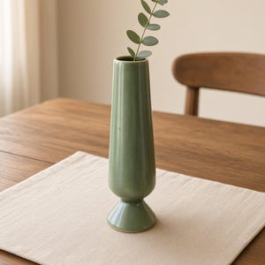 May include: A tall, sage green ceramic vase with a smooth, glossy finish, featuring a slender, tapered body that widens slightly at the base. A single green eucalyptus stem with small, rounded leaves is placed inside the narrow opening.