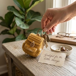 May include: A mustard yellow crocheted miniature backpack keychain featuring a small wooden button closure on the front flap. The backpack is attached to a gold-toned metal keyring with several keys and a small rectangular metal tag attached.