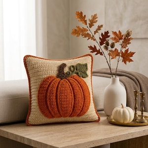 May include: A square crochet throw cushion in a light beige colour featuring a large, textured, three-dimensional orange pumpkin appliqué with a dark brown stem and a green leaf. The cushion has a matching orange crocheted border trim.