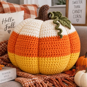May include: A round, crocheted pumpkin-shaped cushion featuring horizontal stripes of white, orange, and yellow yarn. The top includes a dark brown crocheted stem and a textured green crocheted leaf with a curly vine detail attached to the side.