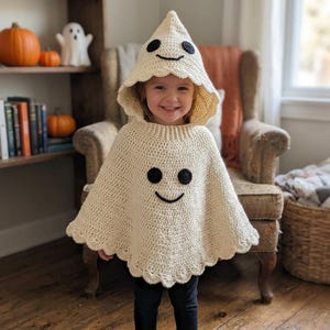 Puede incluir: Un poncho de ganchillo color crema con capucha diseñado para parecer un fantasma. La prenda presenta una capucha puntiaguda, un dobladillo festoneado y dos ojos circulares negros con una boca curva negra cosidos en la parte delantera y la capucha.
