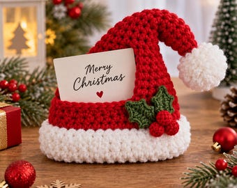 Puede incluir: Un tarjetero de ganchillo con forma de gorro de Papá Noel en rojo y blanco, con un pompón blanco en la punta y una hoja de acebo verde con tres bayas rojas en el lateral. Una tarjeta blanca con el texto Merry Christmas y un pequeño corazón.