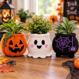 Puede incluir: Tres fundas de ganchillo para macetas con diseños de Halloween. La izquierda es una calabaza naranja con una enredadera verde, el centro es un fantasma blanco con mejillas rosas y la derecha es un caldero negro con telaraña y araña moradas.