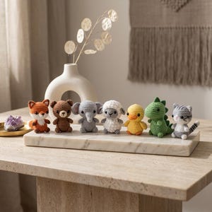 May include: A collection of seven small crocheted animal figures including a fox, bear, elephant, sheep, duck, dinosaur, and cat. Each figure is crafted with yarn in distinct colors and features small black eyes, arranged in a row on a marble slab.