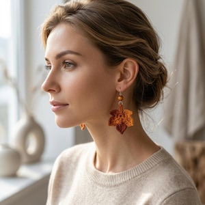 May include: A pair of dangle earrings featuring a crocheted maple leaf in shades of burnt orange and brown. Each leaf is attached to a small, smooth, round wooden bead and a silver-toned metal hook, creating a textured, autumn-inspired accessory.