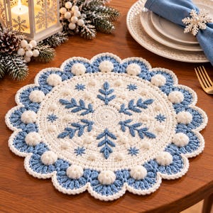 Puede incluir: Un mantel individual redondo de ganchillo con un centro blanco, bordados azules en forma de hoja y pequeñas borlas blancas. El borde consiste en un acabado festoneado azul con borlas blancas, creando un diseño circular.
