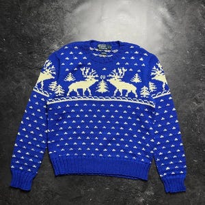 Könnte beinhalten: Ein königsblauer, langärmeliger Strickpullover mit einem cremefarbenen Muster aus Rentieren und Tannenbäumen auf der Brust sowie einem wiederkehrenden kleinen Sternenmotiv auf dem Körper und den Ärmeln. Das Innenetikett zeigt den Text Polo by Ralph Lauren.