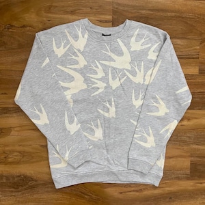 May include: A light grey crewneck sweatshirt featuring a repeating pattern of off-white swallow silhouettes across the front, sleeves, and shoulders. The garment has long sleeves with ribbed cuffs and a ribbed neckline, constructed from a soft fabric.