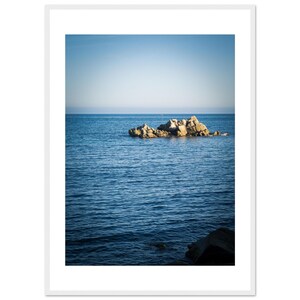 Puede incluir: Una fotografía vertical enmarcada que muestra un grupo de rocas de color marrón claro que emergen del centro de un océano azul profundo bajo un cielo azul claro y despejado. La composición incluye un borde blanco alrededor de la imagen.